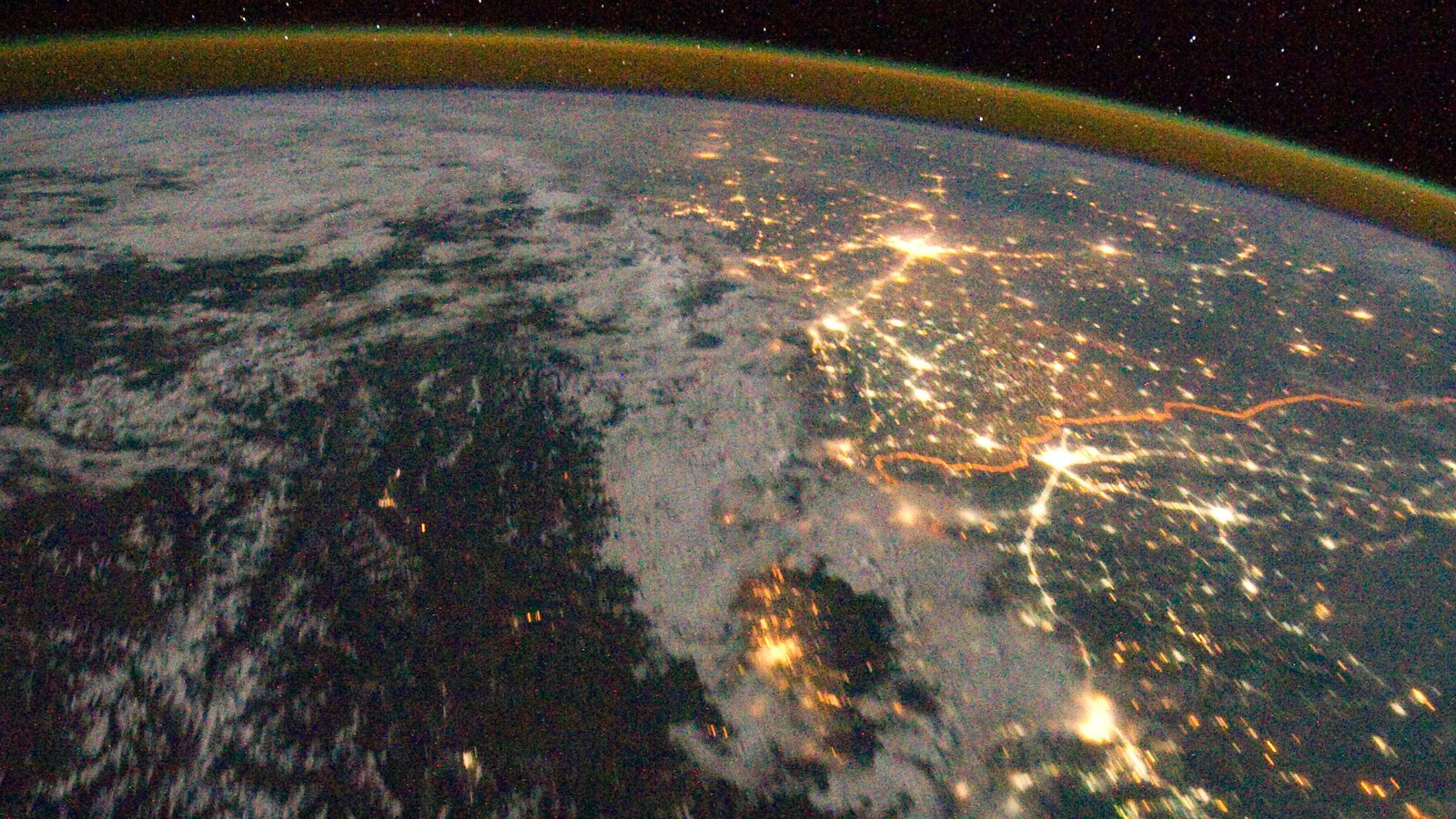 The India-Pakistan border at night from the International Space Station: city lights scattered across the dark land, the floodlit border fence a continuous orange line running through them